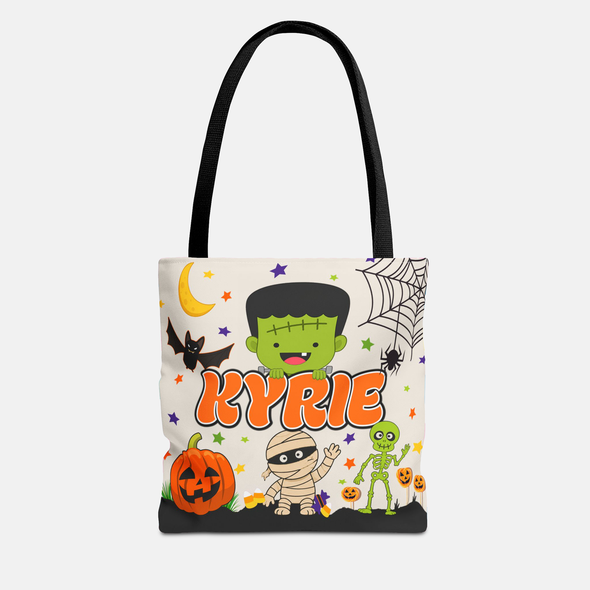 A white canvas tote bag with black shoulder straps, featuring a colorful Halloween-themed cartoon print with the name 'KYRIE' in orange. The design includes Frankenstein's monster, a mummy, a green alien, a jack-o'-lantern, a spiderweb, and stars on a light gray background.