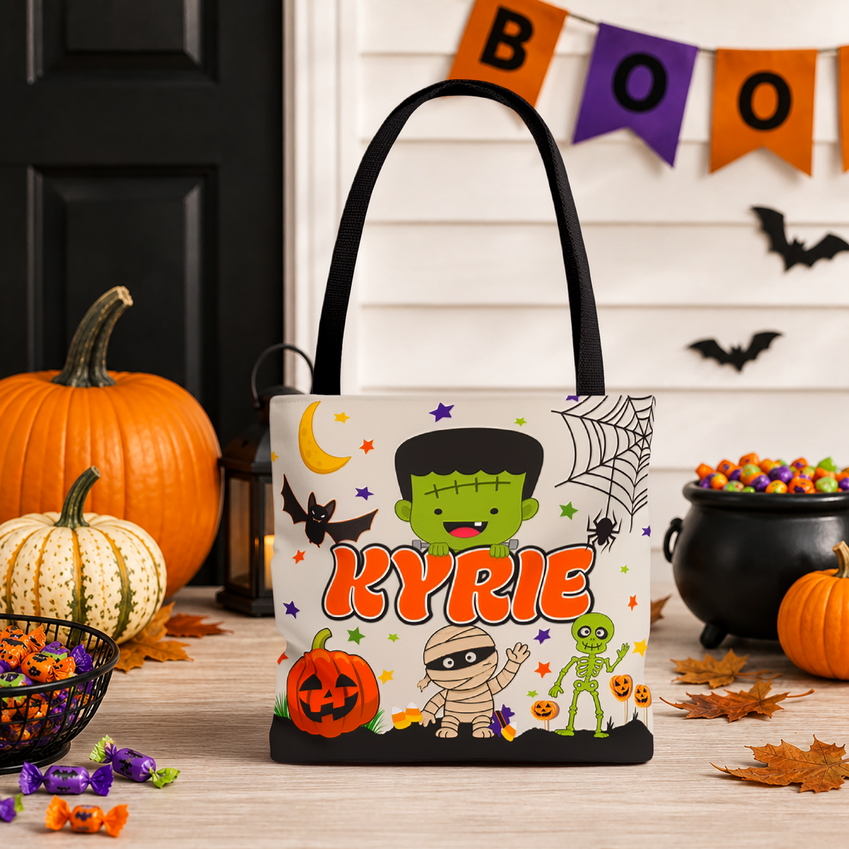 A Halloween-themed tote bag with black handles stands on a wooden table in a decorated fall setting. The bag features cartoon characters including Frankenstein's monster, a mummy, a green alien, a pumpkin, and spider.