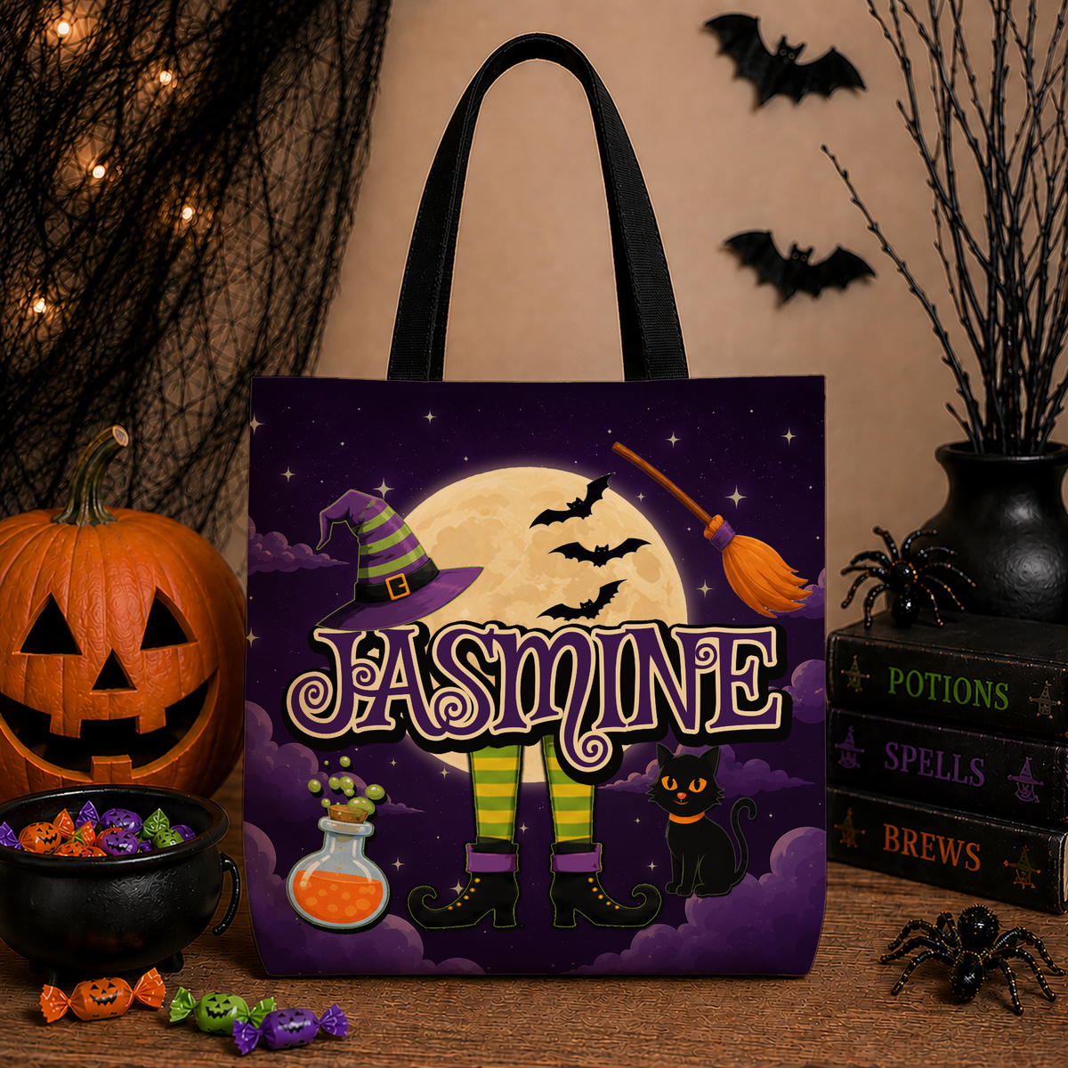 A Halloween-themed tote bag with black handles, featuring a purple and yellow moonlit background with the name 'JASMINE' in large decorative letters, a witch's hat, broom, bats, boots, and a black cat. The bag is styled on a tabletop with pumpkins, candy, books, and spider decorations.