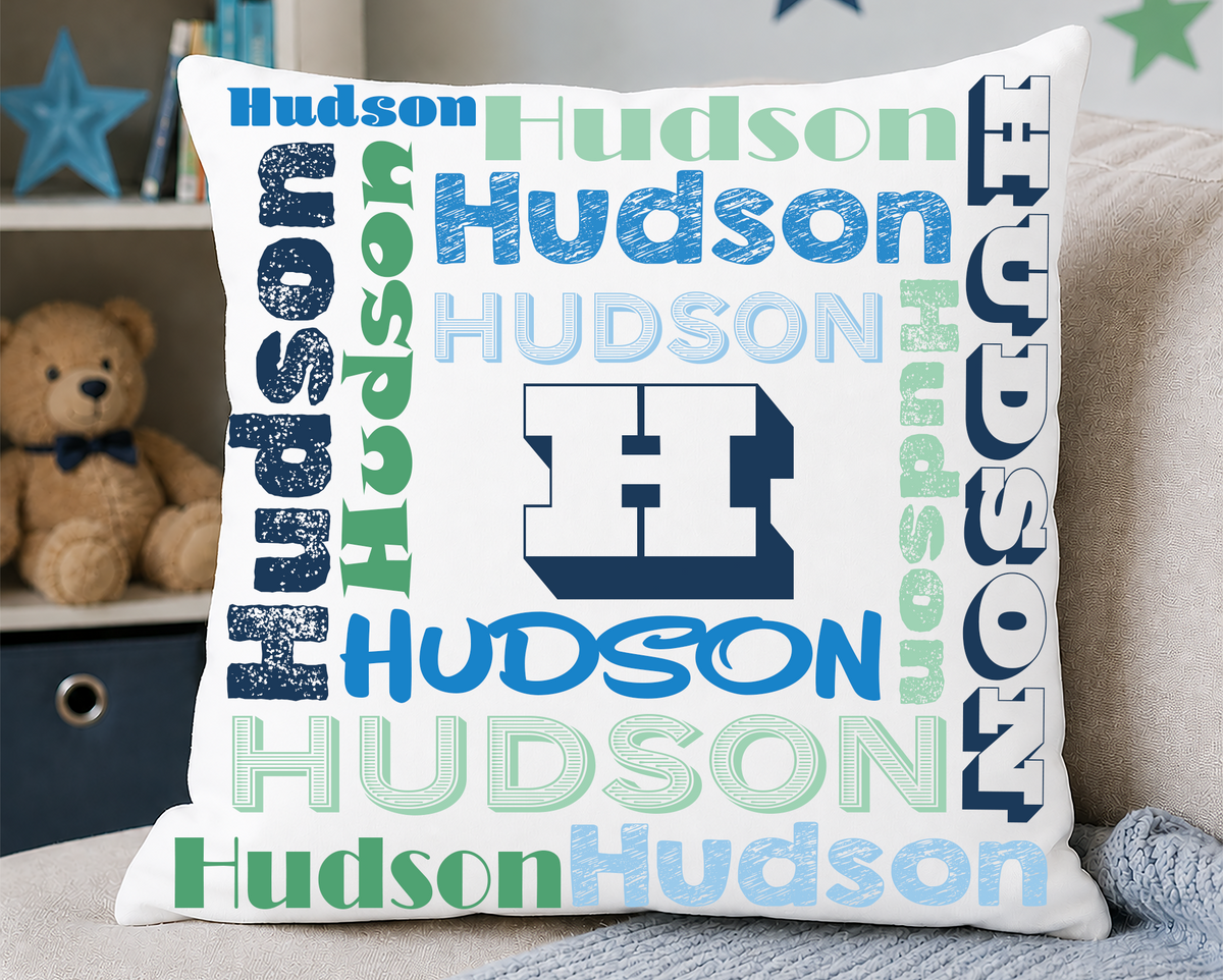 A square white throw pillow featuring a colorful 'Hudson' word cloud design with a large block 'H' logo in the center, styled on a beige couch with a gray blanket and a teddy bear in the background.