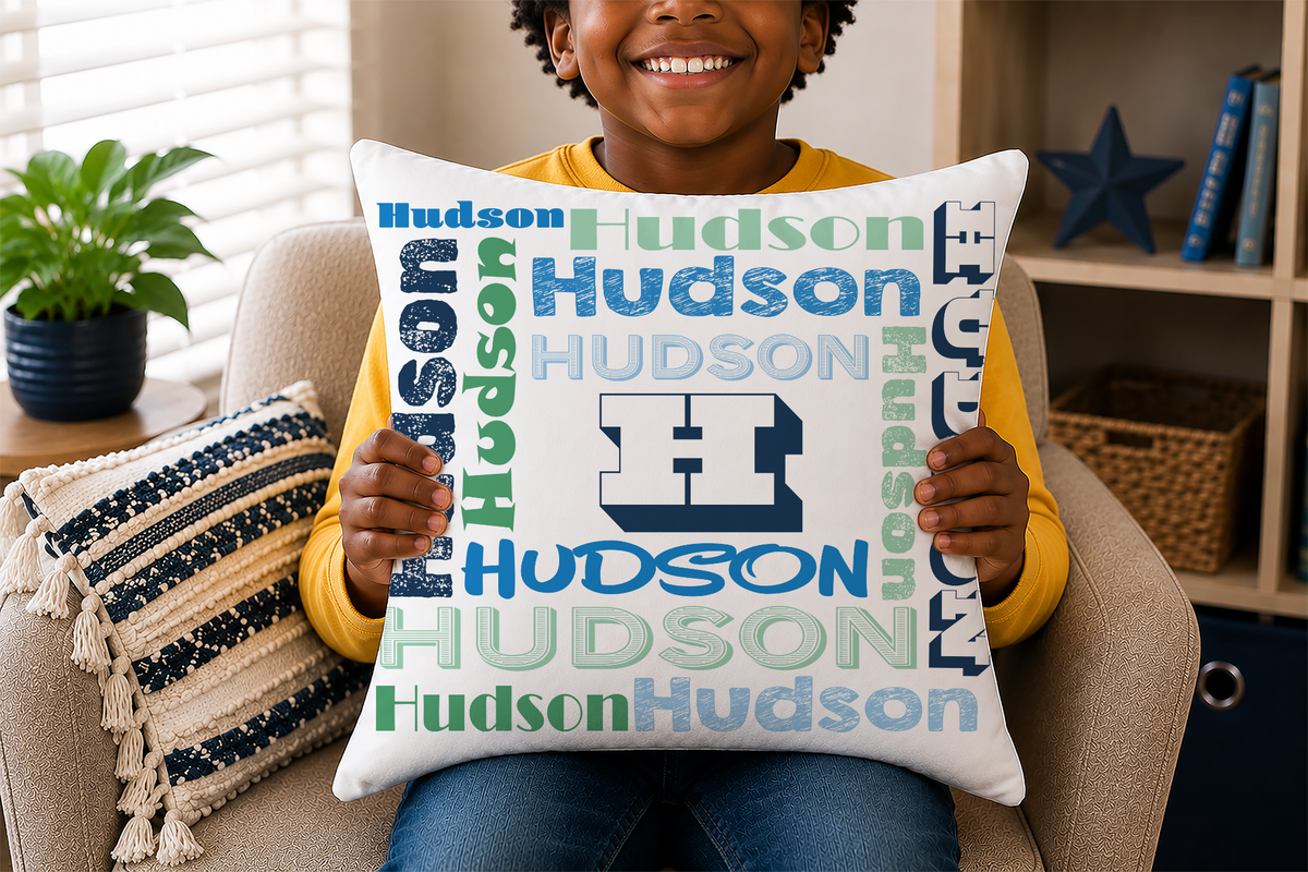 A smiling young child seated in a beige armchair holds a square throw pillow printed with repeated 'Hudson' text in blue and green tones and a large dark blue 'H' logo. The room includes a plant, shelving, and a striped accent pillow.