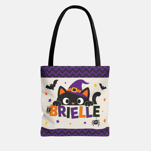 A white canvas tote bag with black handles, featuring a Halloween-themed graphic with a black cat in a purple witch hat, bats, spiders, stars, and the name 'BRIELLE' in colorful letters. The bag has purple chevron borders at the top and bottom.
