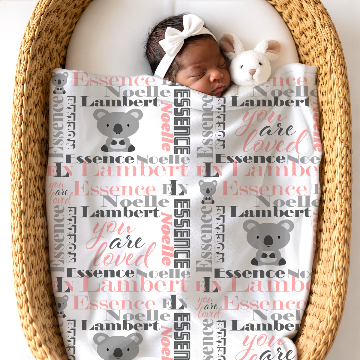 A newborn baby sleeping peacefully in a woven bassinet, wrapped in a white personalized blanket with koala graphics and the name 'Essence Noelle Lambert', wearing a matching white bow headband and holding a plush bunny.