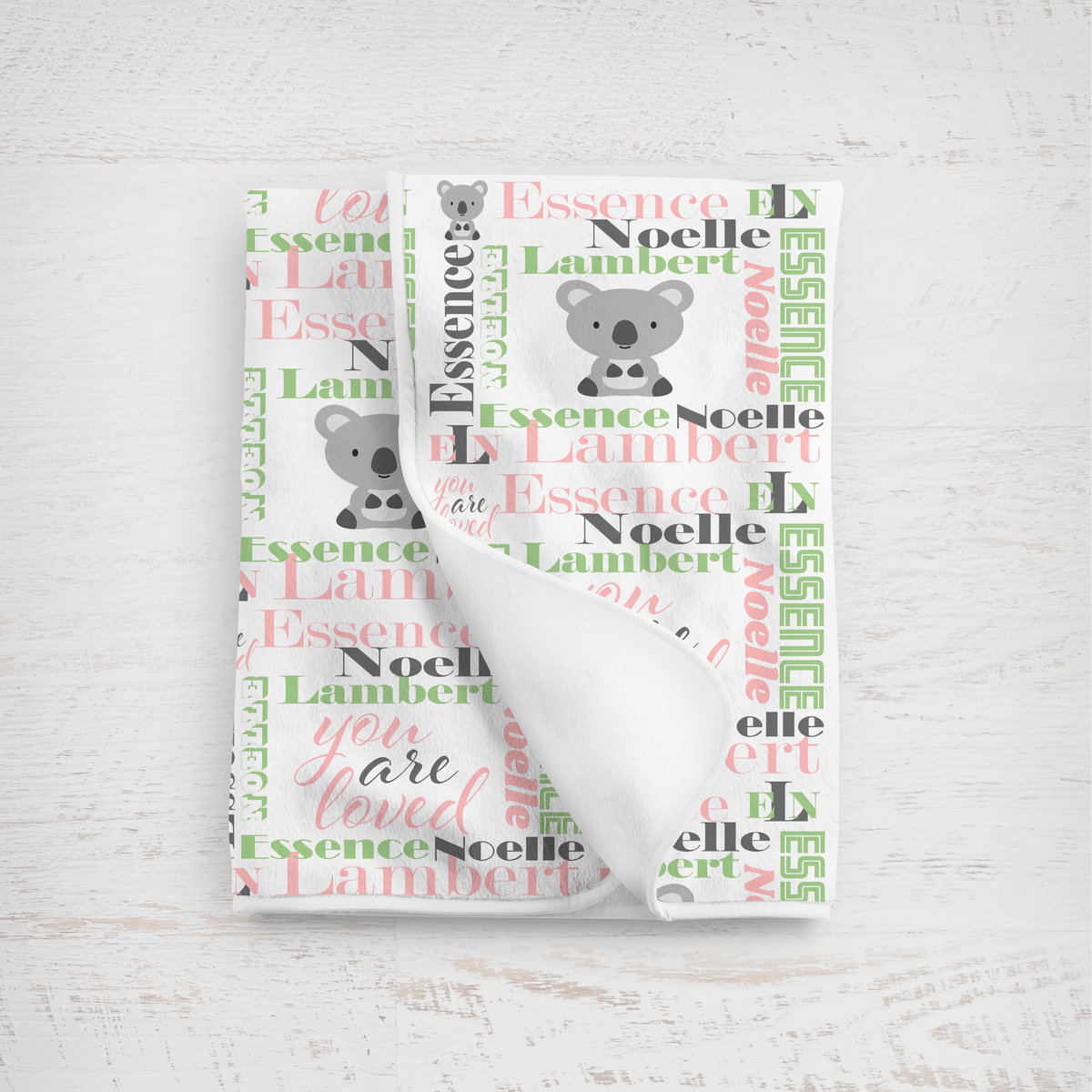 A folded white baby blanket with a personalized koala print, featuring the name 'Essence Noelle Lambert' and the phrase 'you are loved' in pink and green apple script.
