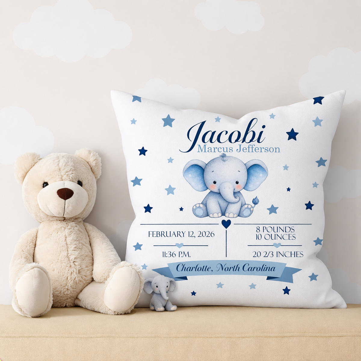 A personalized baby birth announcement pillow displayed next to a beige teddy bear and a small plush elephant. The pillow is white with blue stars, a blue elephant illustration, and custom text reading 'Jacobi Marcus Jefferson' with birth details and a banner reading 'Charlotte, North Carolina'.