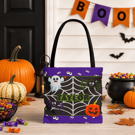 A Halloween-themed tote bag with black handles stands on a tabletop decorated with pumpkins, candy, a cauldron, bats, and a BOO banner. The bag features a purple top and bottom border, a black center with a spiderweb pattern, cartoon ghosts, spiders, and a jack-o'-lantern, with the name 'TAYLOR' printed in green across the middle.