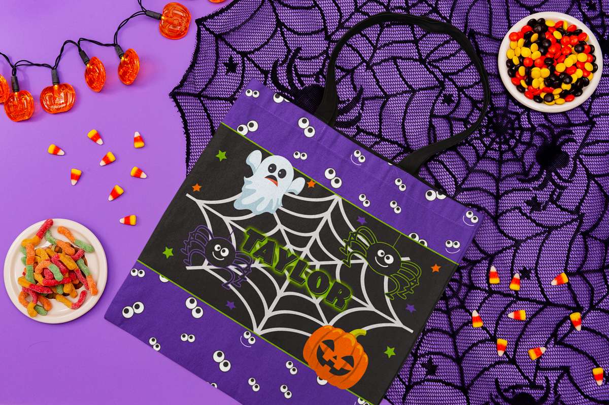 A purple Halloween-themed reusable tote bag with black handles, featuring a black central panel decorated with a spiderweb, cartoon ghosts, a jack-o'-lantern, and the name 'TAYLOR' in green. The bag is styled in a flat-lay arrangement on a purple background with spiderwebbing, candy corn, gummy candies, and orange string lights.