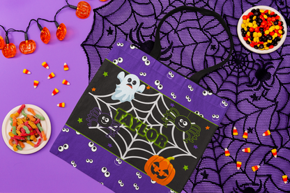 A purple Halloween-themed reusable tote bag with black handles, featuring a black central panel decorated with a spiderweb, cartoon ghosts, a jack-o'-lantern, and the name 'TAYLOR' in green. The bag is styled in a flat-lay arrangement on a purple background with spiderwebbing, candy corn, gummy candies, and orange string lights.