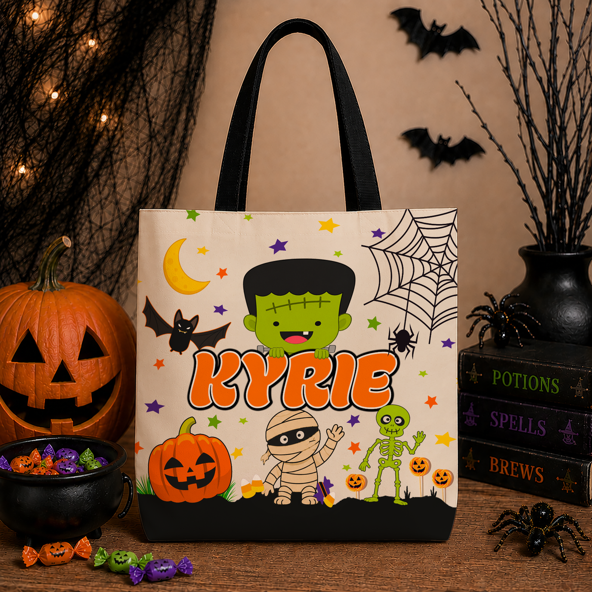 A personalized Halloween tote bag with a Frankenstein-themed design, featuring the name 'KYRIE' in orange. The bag is styled on a wooden table with a pumpkin, candy bowl, spider decorations, and books in the background.