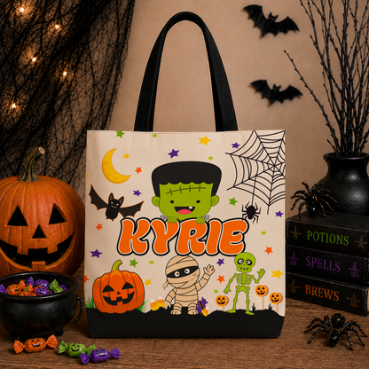 A personalized Halloween tote bag with a Frankenstein-themed design, featuring the name 'KYRIE' in orange. The bag is styled on a wooden table with a pumpkin, candy bowl, spider decorations, and books in the background.