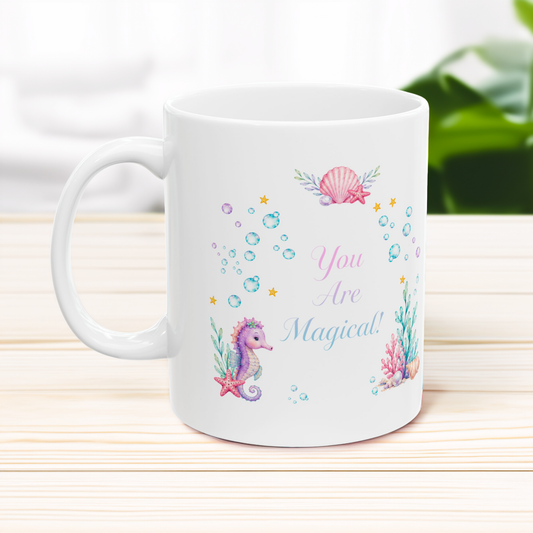 A white ceramic mug with a colorful underwater theme print featuring a seahorse, seashell, bubbles, and the text 'You Are Magical!' in pink and blue lettering, set on a light wooden surface with a blurred background.
