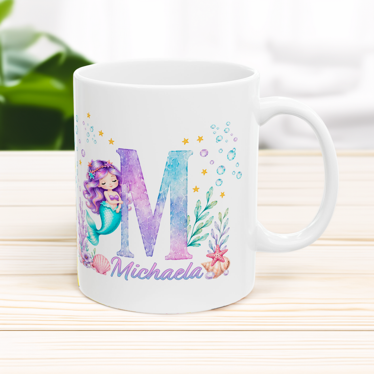 A white ceramic mug with a colorful mermaid-themed personalized design, featuring a large letter 'M', an underwater mermaid illustration, bubbles, and the name 'Michaela' in purple script, set on a light wooden surface.