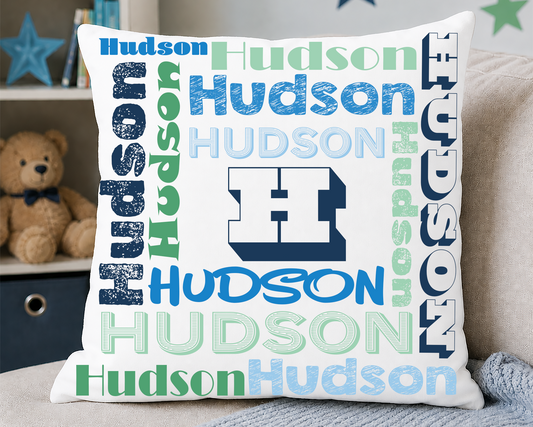 A square white throw pillow featuring a colorful 'Hudson' word cloud design with a large block 'H' logo in the center, styled on a beige couch with a gray blanket and a teddy bear in the background.