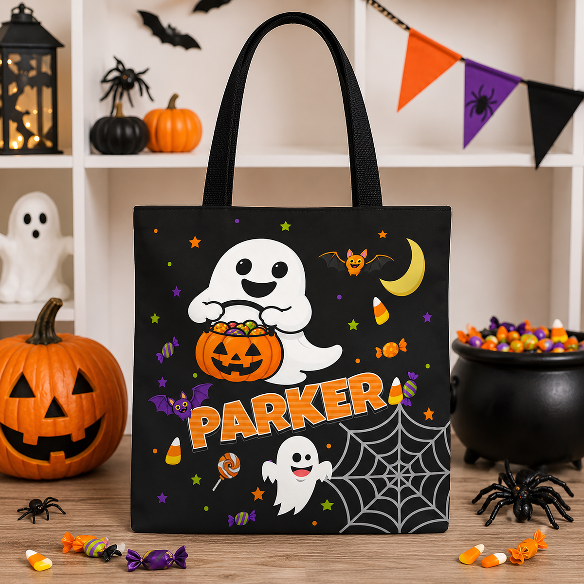 A black Halloween tote bag with a custom printed design featuring a white cartoon ghost holding an orange pumpkin bucket, the name 'PARKER' in orange, a crescent moon, bats, candy corn, spiders, and spiderwebs. The bag is displayed on a wooden surface surrounded by Halloween decorations including a jack-o'-lantern, a cauldron of candy, and a black spider.