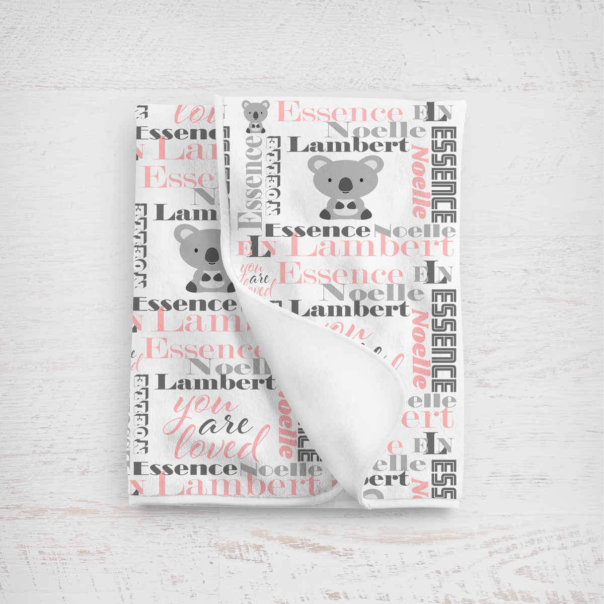 A folded white baby blanket with a personalized koala print, featuring the name 'Essence Noelle Lambert' in charcoal gray and pink script alongside koala illustrations, displayed on a light wood background.