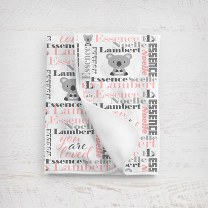 A folded white baby blanket with a personalized koala print, featuring the name 'Essence Noelle Lambert' in charcoal gray and pink script alongside koala illustrations, displayed on a light wood background.