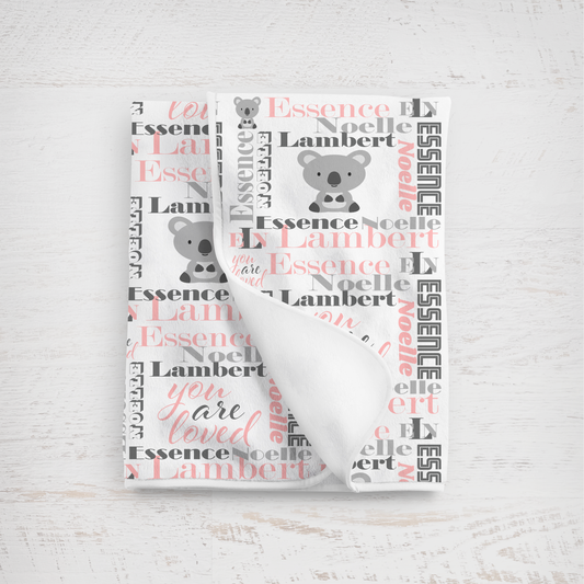 A folded white baby blanket with a personalized koala print, featuring the name 'Essence Noelle Lambert' in charcoal gray and pink script alongside koala illustrations, displayed on a light wood background.
