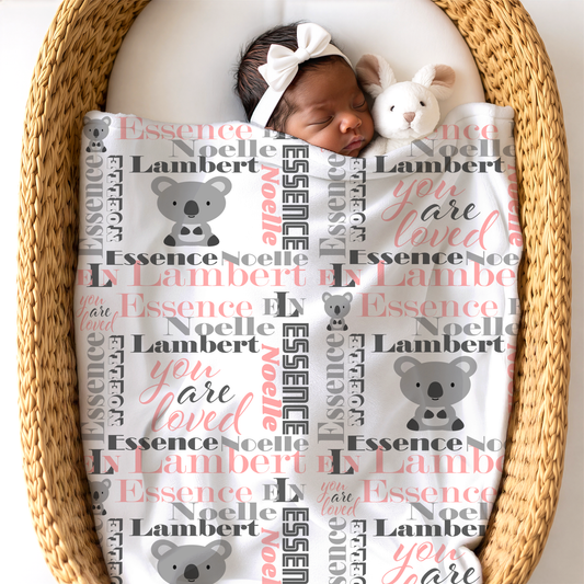 A newborn baby sleeping peacefully in a woven bassinet, wrapped in a white personalized blanket with koala graphics and the name 'Essence Noelle Lambert', wearing a matching white bow headband and holding a plush bunny.