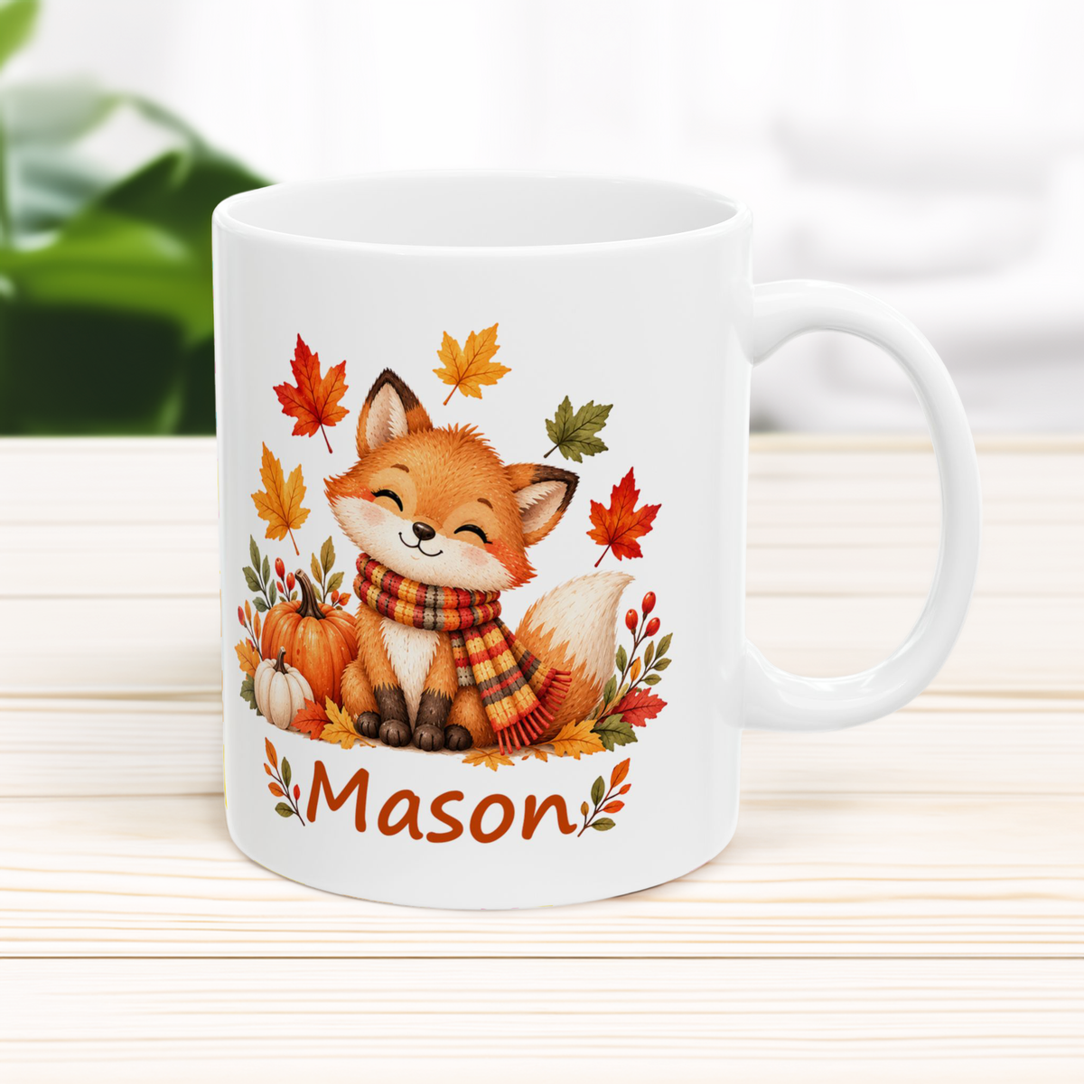 A white ceramic mug with a custom illustration of a smiling orange fox wearing a plaid scarf, surrounded by autumn leaves and pumpkins, with the name 'Mason' printed below. The mug is set on a light wood surface with a soft-focus background.