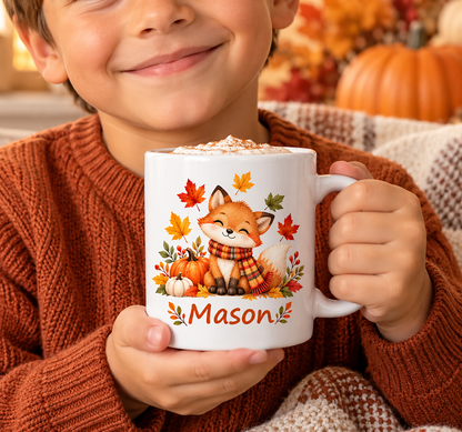 A child in a rust-orange knit sweater holds a white ceramic mug with a fall-themed cartoon fox design and the name Mason printed on it. The mug is filled with a hot beverage topped with whipped cream, with a cozy autumn background featuring pumpkins and a blanket.