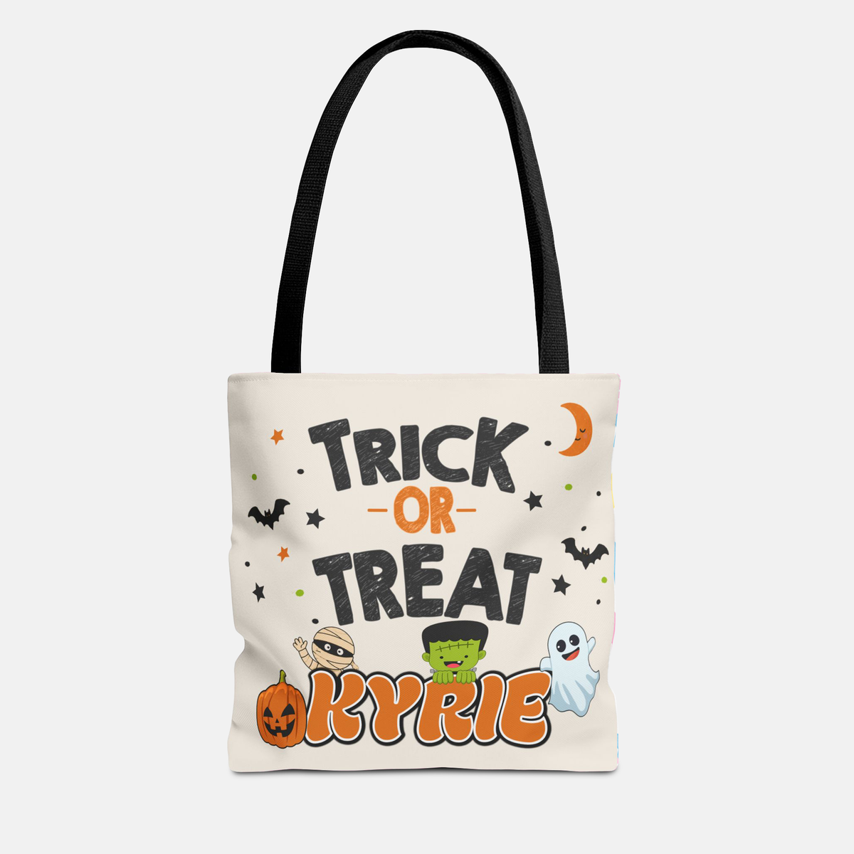 An off-white canvas tote bag with black shoulder straps, featuring a Halloween-themed graphic print that reads 'TRICK OR TREAT KYRIE' with cartoon pumpkins, ghosts, and bats on a light gray background.