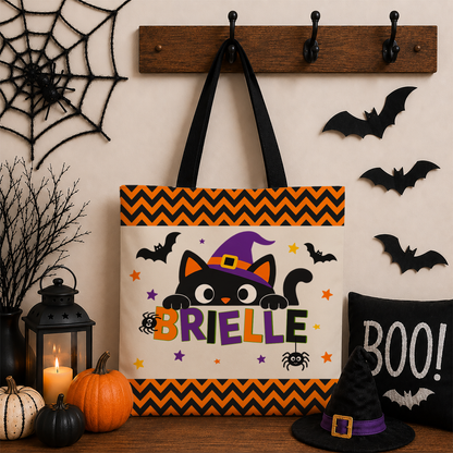 A Halloween-themed tote bag hanging on a wall hook, decorated with a black cat in a purple witch hat, the name 'BRIELLE', bats, spiders, and orange chevron borders. The bag is styled with pumpkins, a lantern, a black 'BOO!' pillow, and a witch hat against a beige wall with bat cutouts.