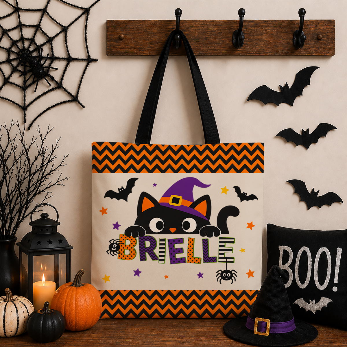 A personalized Halloween tote bag hanging on a wooden wall hook, surrounded by seasonal props including a pumpkin lantern, black spider web, bat cutouts, a witch hat, and a black pillow that says 'BOO!'. The bag features a black cat wearing a purple witch hat peeking behind the text 'BRIELLE' with orange and black chevron borders.