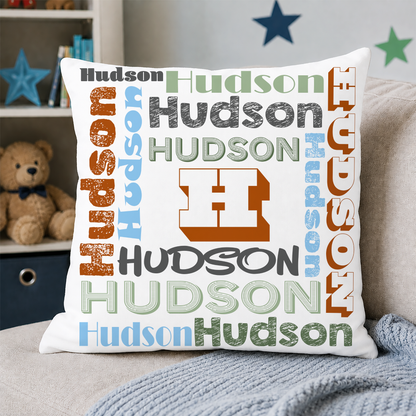 A square white throw pillow featuring a colorful 'Hudson' wordcloud design with a large central orange block 'H', styled on a gray sofa with a knitted blanket and a teddy bear in the background.
