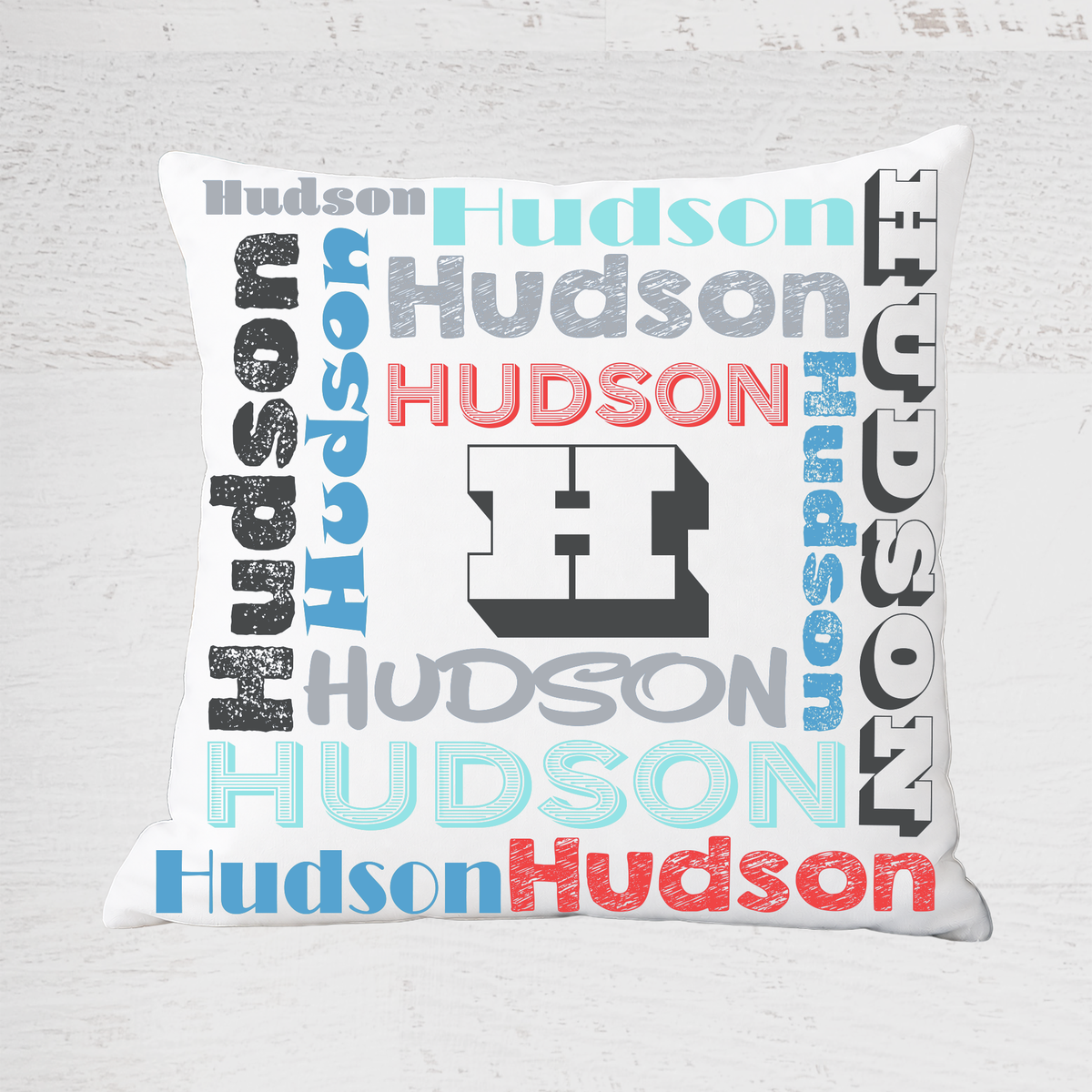 A square white throw pillow featuring a 'Hudson' wordcloud design with various stylized fonts and colors, including a large central block 'H' logo, set against a light gray wood-textured background.
