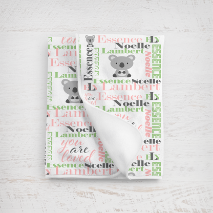 A folded white baby blanket with a personalized koala print, featuring the name  'Essence Noelle Lambert' and the phrase 'you are loved' in pink and green apple script.