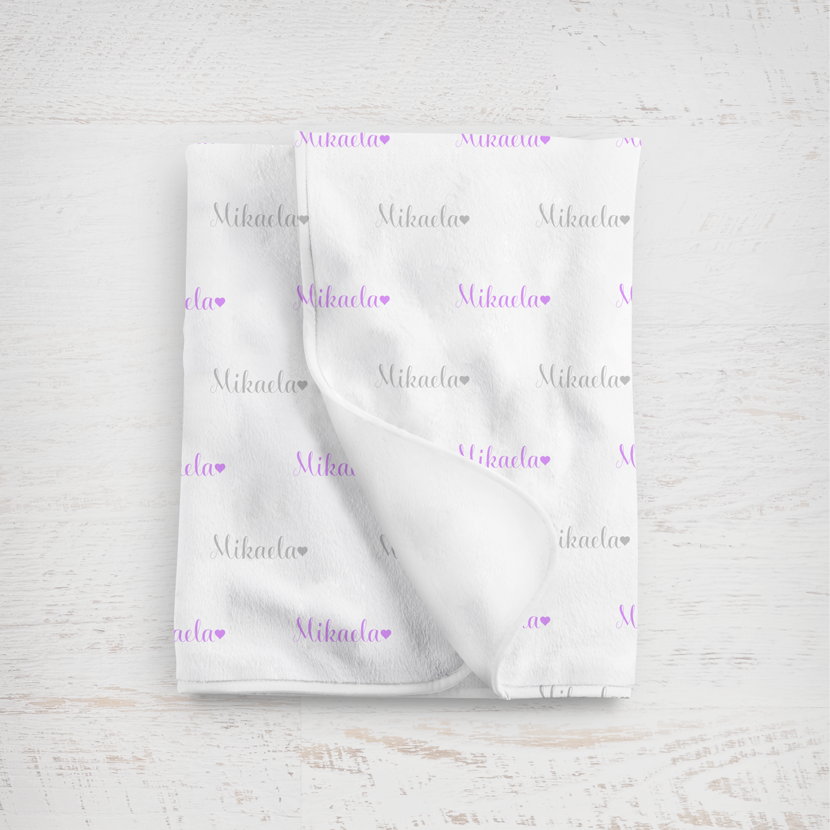 White blanket with name in lavender and gray printed repeatedly on a light wooden surface