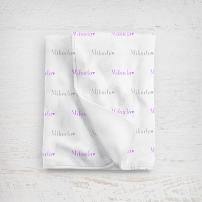 White blanket with name in lavender and gray printed repeatedly on a light wooden surface