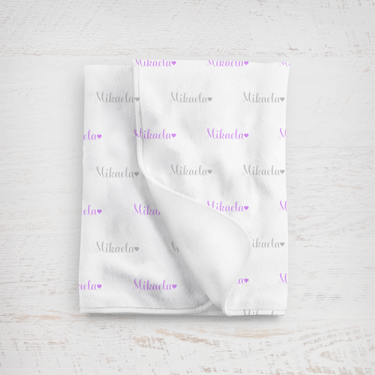 White blanket with name in lavender and gray printed repeatedly on a light wooden surface