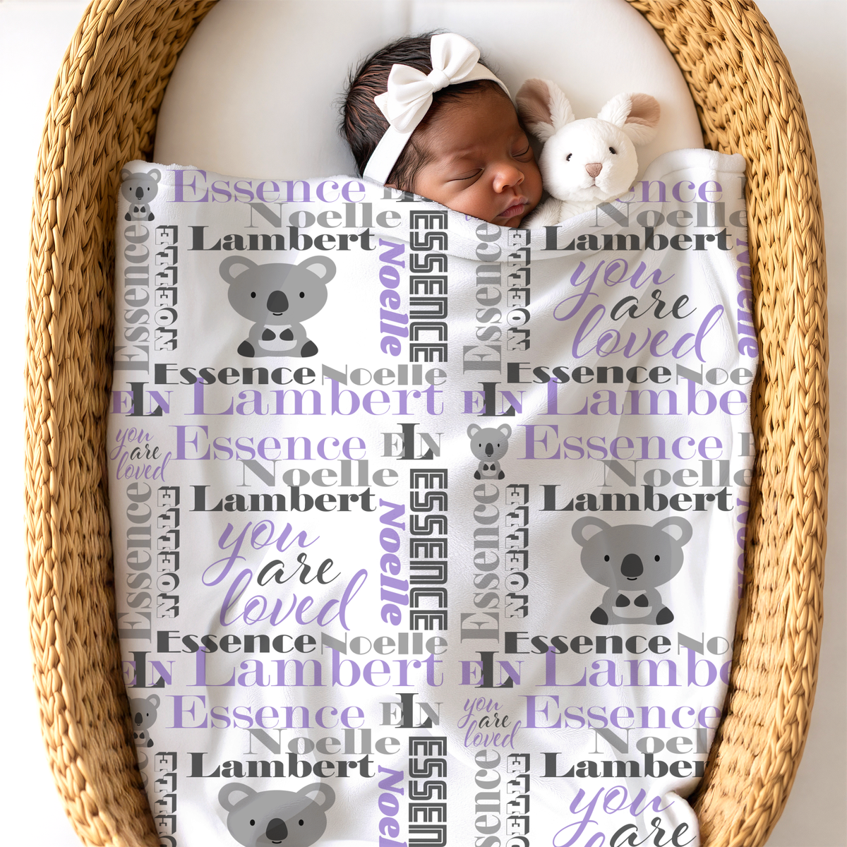 A sleeping newborn baby nestled in a woven wicker bassinet, wrapped in a white blanket printed with grey koalas, lavender text reading 'you are loved', and charcoal gray and lavender personalized name 'Essence Noelle Lambert'. The baby wears a white bow headband and has a small plush bunny toy.