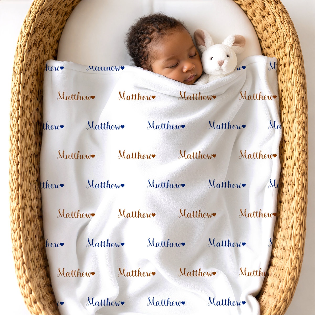 A sleeping newborn baby wrapped in a personalized white blanket printed with the name 'Matthew' and small hearts, resting inside a woven wicker basket next to a white plush bunny toy.